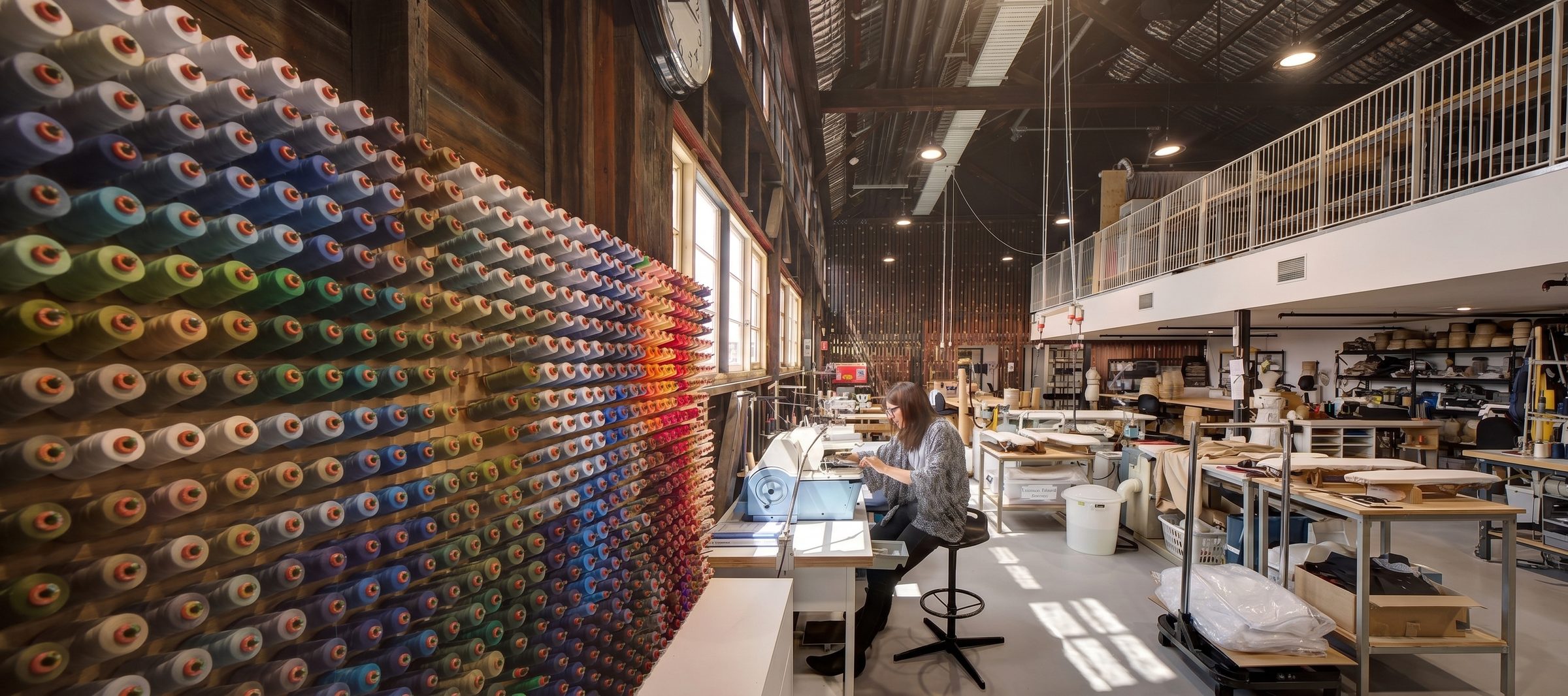 A long workshop floor with a wall of thread spools, work benches and someone working at a machine.
